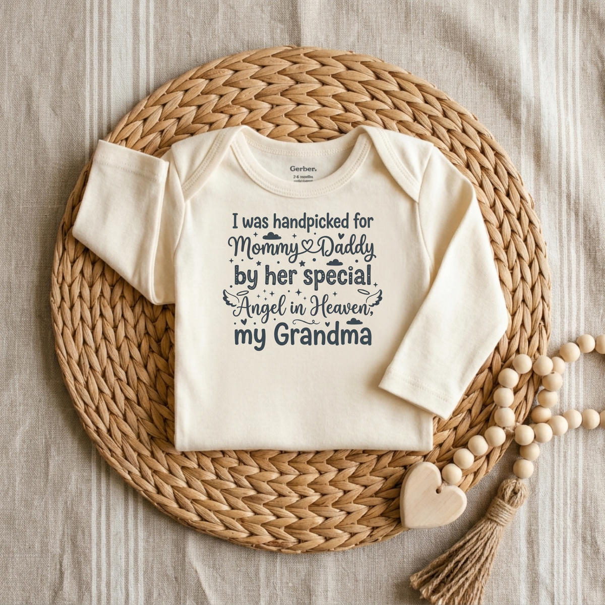 A cream-colored Handpicked by My Angel Grandma Onesie – Memorial Baby Bodysuit from Toodleroos rests on a woven placemat, featuring the touching message “I was handpicked for Mommy & Daddy by her special Angel in Heaven, my Grandma.” Beside this heartfelt angel grandma bodysuit lies a beaded heart-shaped decoration.