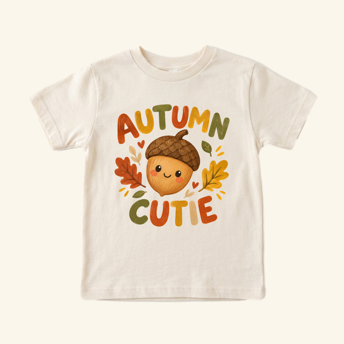 Cream-colored Autumn Cutie Acorn Toddler Shirt with a graphic of a smiling acorn surrounded by autumn leaves and the text Autumn Cutie.
