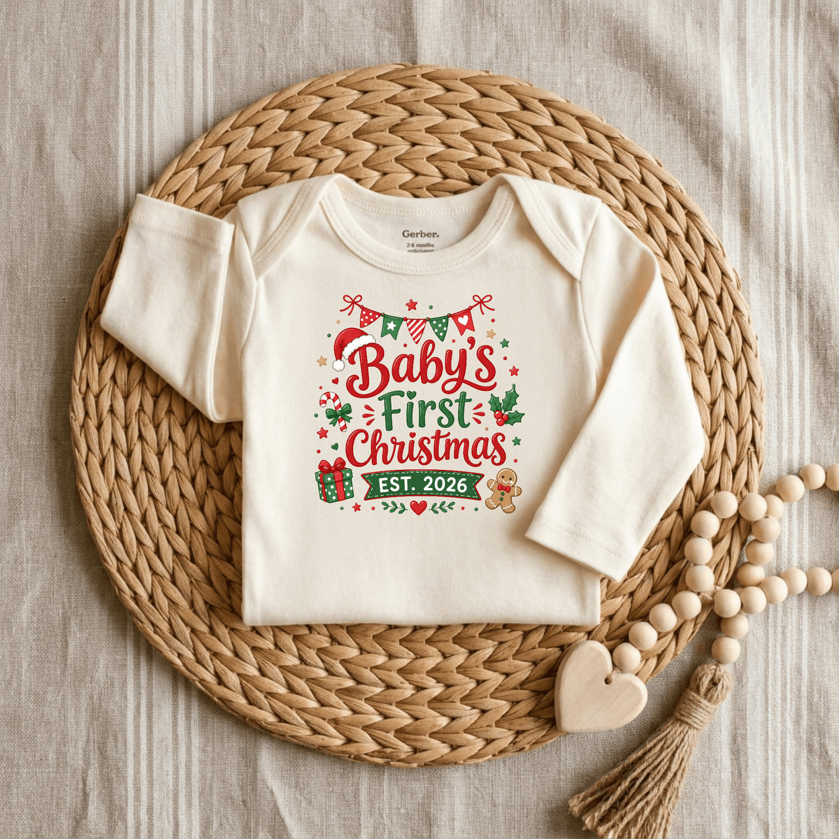 Dressed in a cream Baby’s First Christmas Est. 2026 Long Sleeve Onesie by Toodleroos, this festive holiday keepsake bodysuit features cheerful red and green lettering surrounded by charming seasonal icons. Displayed on a woven placemat with wooden beads and a heart-shaped accent, it’s the perfect 2026 Christmas outfit for your little one’s first celebration.