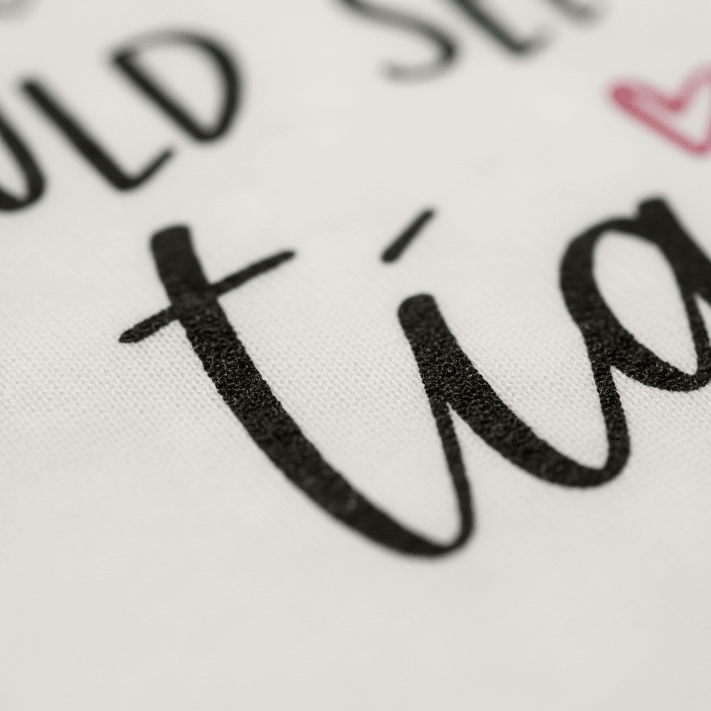 Close-up of black cursive text on white fabric, featuring part of the word “tía” with a pink heart above the letter i—an adorable detail from the Toodleroos “If You Think I’m Cute See My Tía” Toddler Shirt, a funny bilingual tee that makes a sweet aunt gift.