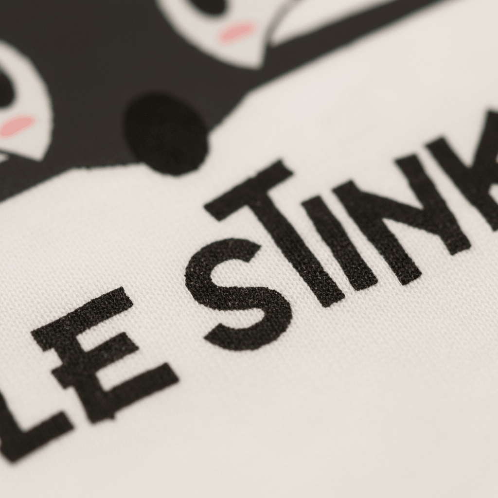 Close-up of black text reading “LE STINK” on white fabric, with part of a cartoon skunk’s face featuring black and pink details visible at the top of the image. This playful Toodleroos Little Stinker Baby Onesie – Funny Skunk Bodysuit makes a charming baby bodysuit and a fun baby shower gift.