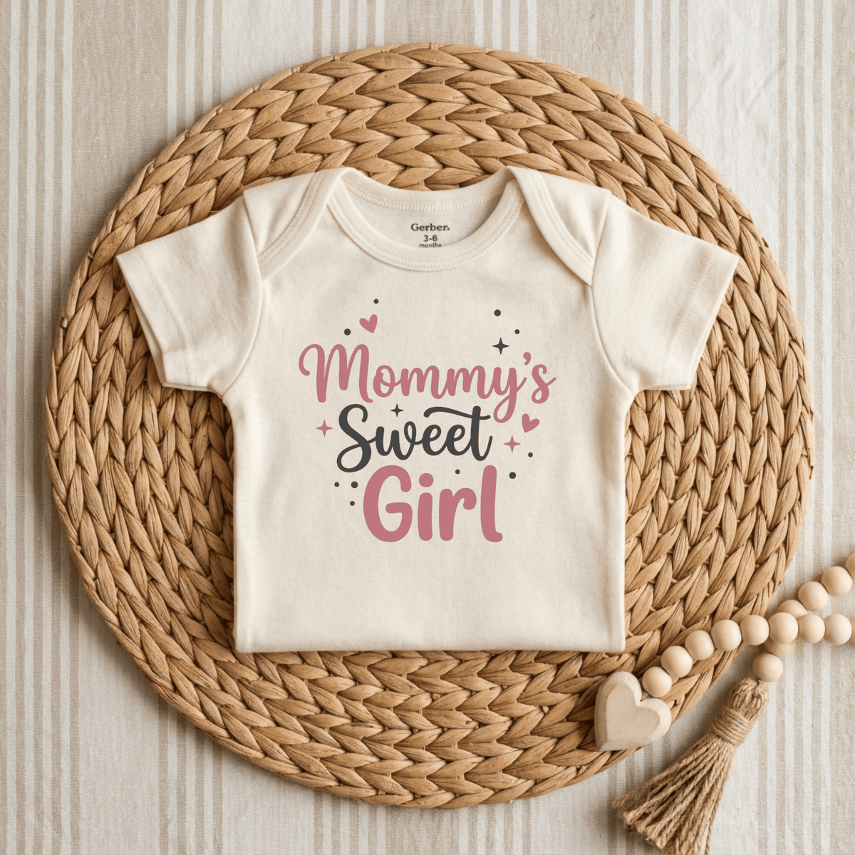 A cream Toodleroos Mommy’s Sweet Girl Onesie – Baby Girl Long Sleeve Bodysuit is displayed on a woven mat, featuring “Mommy’s Sweet Girl” in pink and black letters with heart and sparkle accents. Wooden beads and a heart accessory rest beside this adorable newborn coming-home outfit.