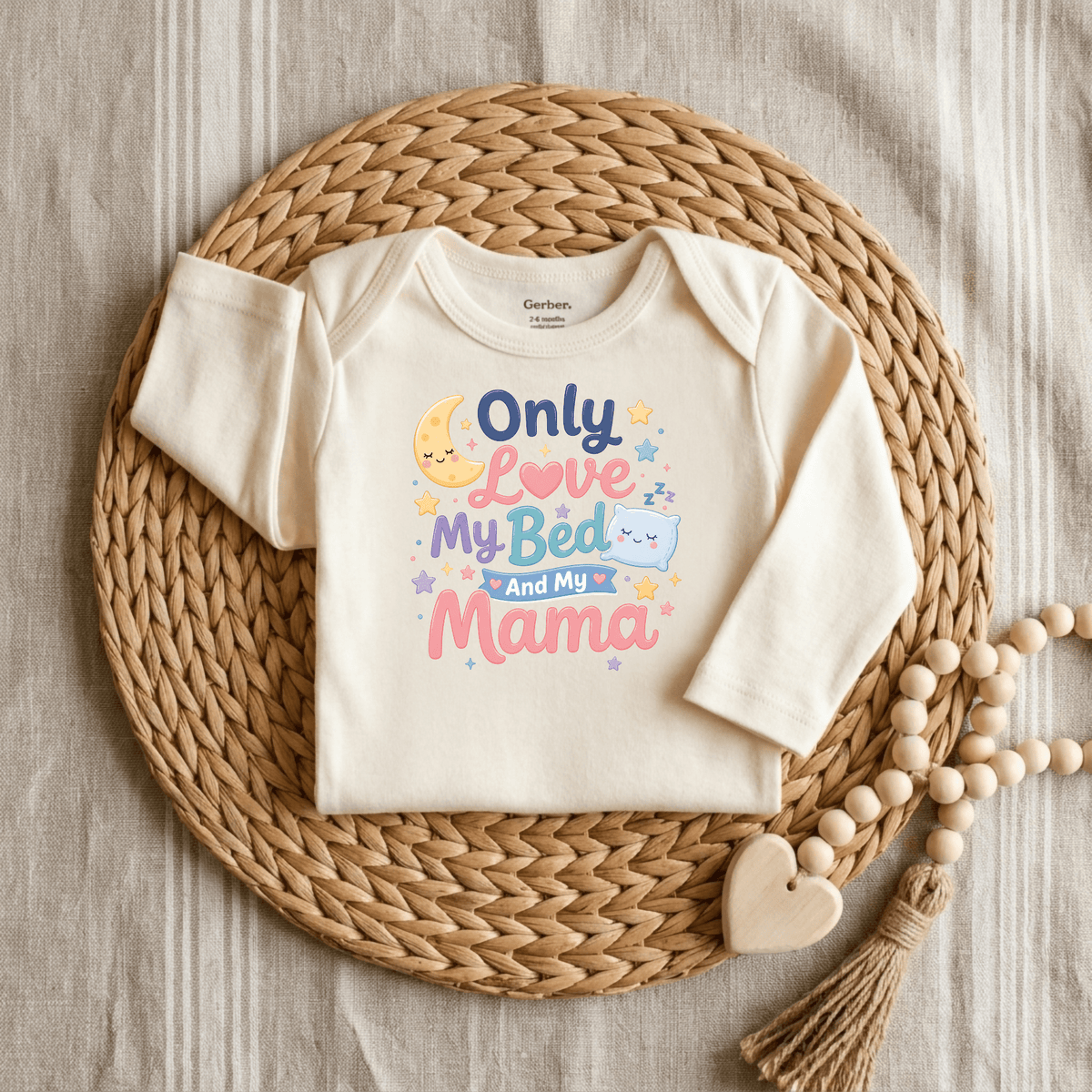 A cream-colored Only Love My Bed and My Mama Onesie – Cute Baby Bodysuit by Toodleroos, featuring colorful text and displayed on a woven placemat beside a beaded wooden heart teether—an adorable, funny, and gender-neutral baby gift.