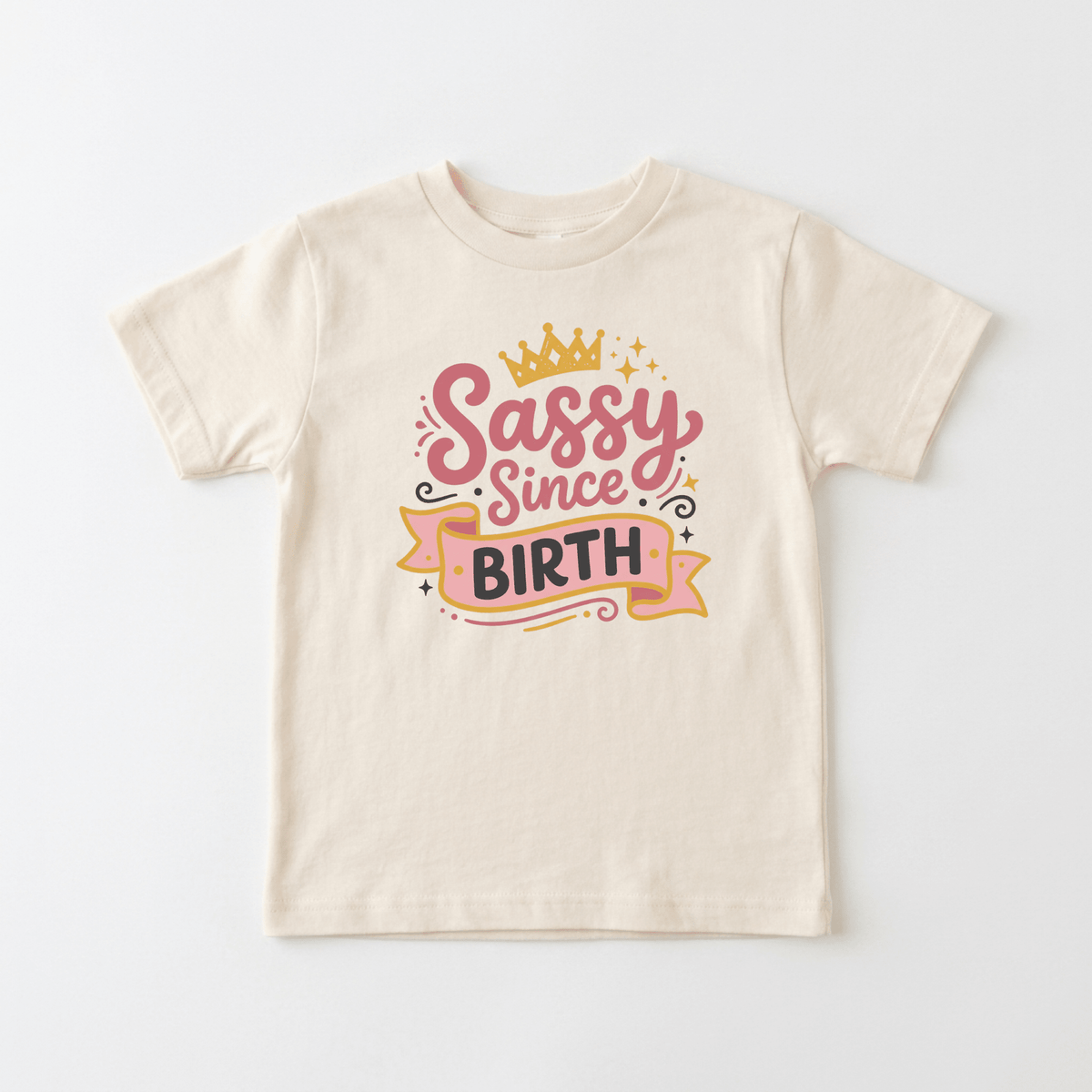 Outfit your little one in the Toodleroos Sassy Since Birth Toddler Tee – a funny girls shirt featuring playful pink and black lettering on beige, accented with a yellow crown, stars, and swirls—perfect for toddlers with big personalities.