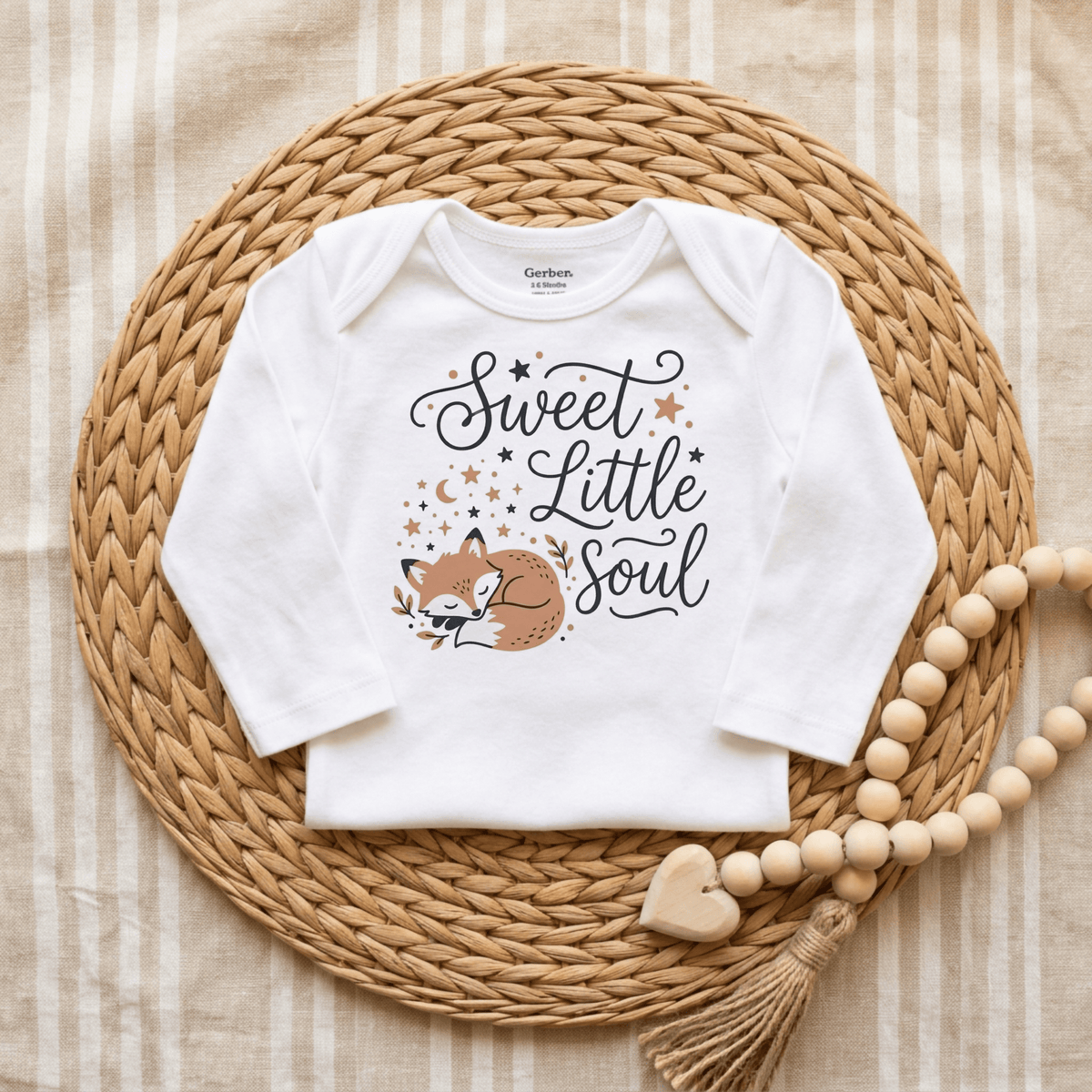 The Sweet Little Soul Onesie – Cute Sleeping Fox Bodysuit by Toodleroos features an adorable sleeping fox and the phrase “Sweet Little Soul.” Displayed on a round woven mat with wooden beads and a heart atop striped fabric, this gender-neutral bodysuit is perfect for any baby.