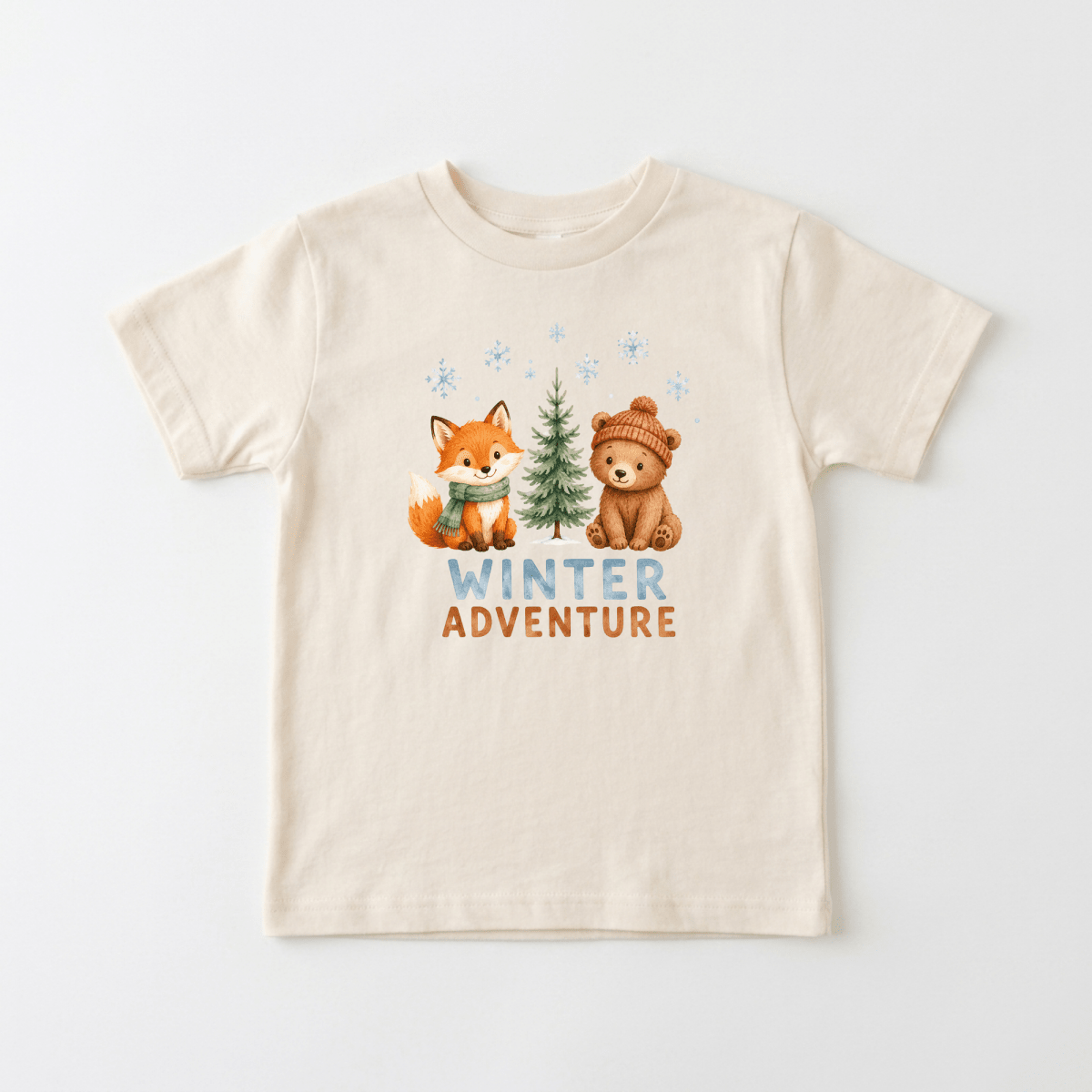 The Winter Adventure Toddler Shirt – Woodland Animal Design by Toodleroos is a beige t-shirt featuring an illustrated fox and bear, along with a pine tree and snowflakes. “WINTER ADVENTURE” is printed below the animals in blue and brown letters, making it the perfect winter shirt for toddlers who love woodland adventures.