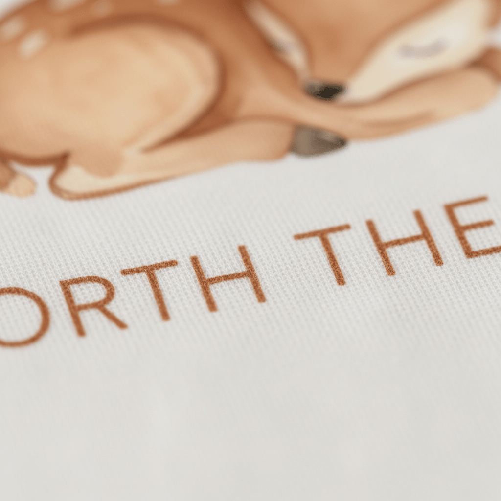 A close-up of a fabric featuring an illustration of a sleeping deer and partially visible brown text that reads “WORTH THE,” from the Toodleroos Worth the Wait Baby Onesie – Sleeping Deer Bodysuit, perfect as an adorable woodland baby shower gift.