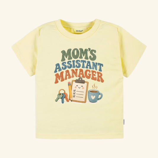 Pale yellow Mom’s Assistant Manager Toddler Shirt featuring a retro design with Moms Assistant Manager text above an illustration of a smiley-faced clipboard, pencil, key ring, and coffee mug.