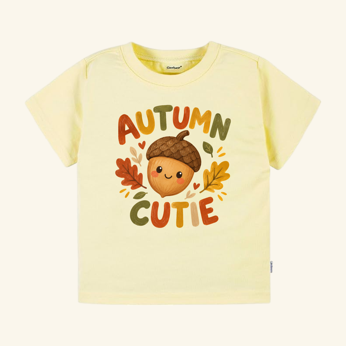 Light yellow Autumn Cutie Acorn Toddler Shirt featuring a smiling cartoon acorn surrounded by colorful leaves and the text AUTUMN CUTIE.