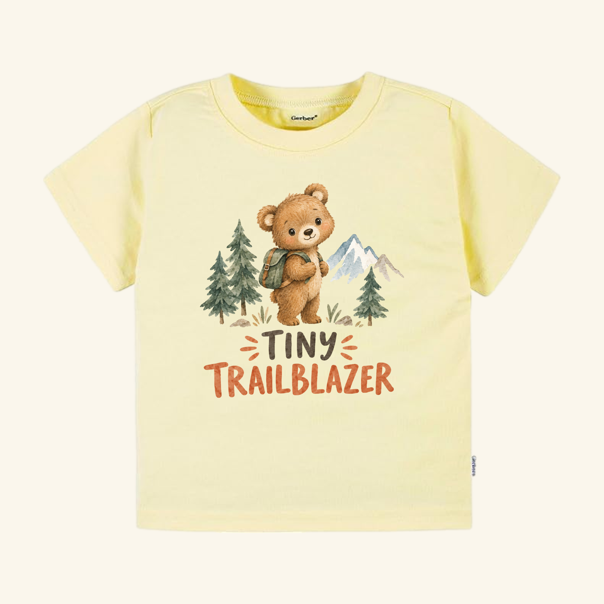 Pale yellow Toodleroos Tiny Trailblazer Toddler Shirt – Hiking Bear Adventure Tee featuring a watercolor illustration of a bear with a backpack among pine trees and mountains, with TINY TRAILBLAZER text underneath.