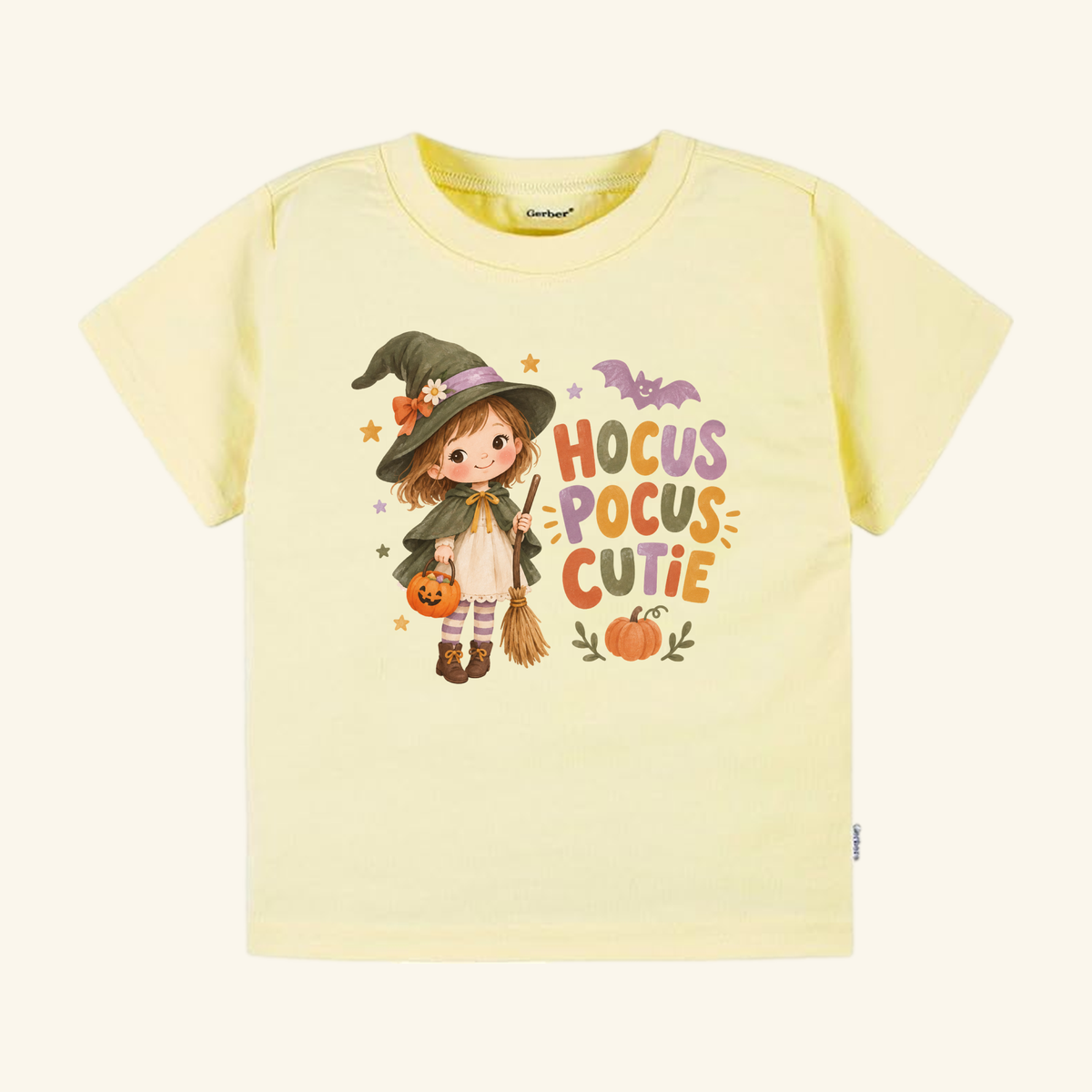 Toodleroos Hocus Pocus Cutie Witch Toddler Halloween Shirt: a pale yellow toddler tee featuring a girl in a witch costume holding a pumpkin bucket and broom, accompanied by a bat, a pumpkin, and the text Hocus Pocus Cutie.