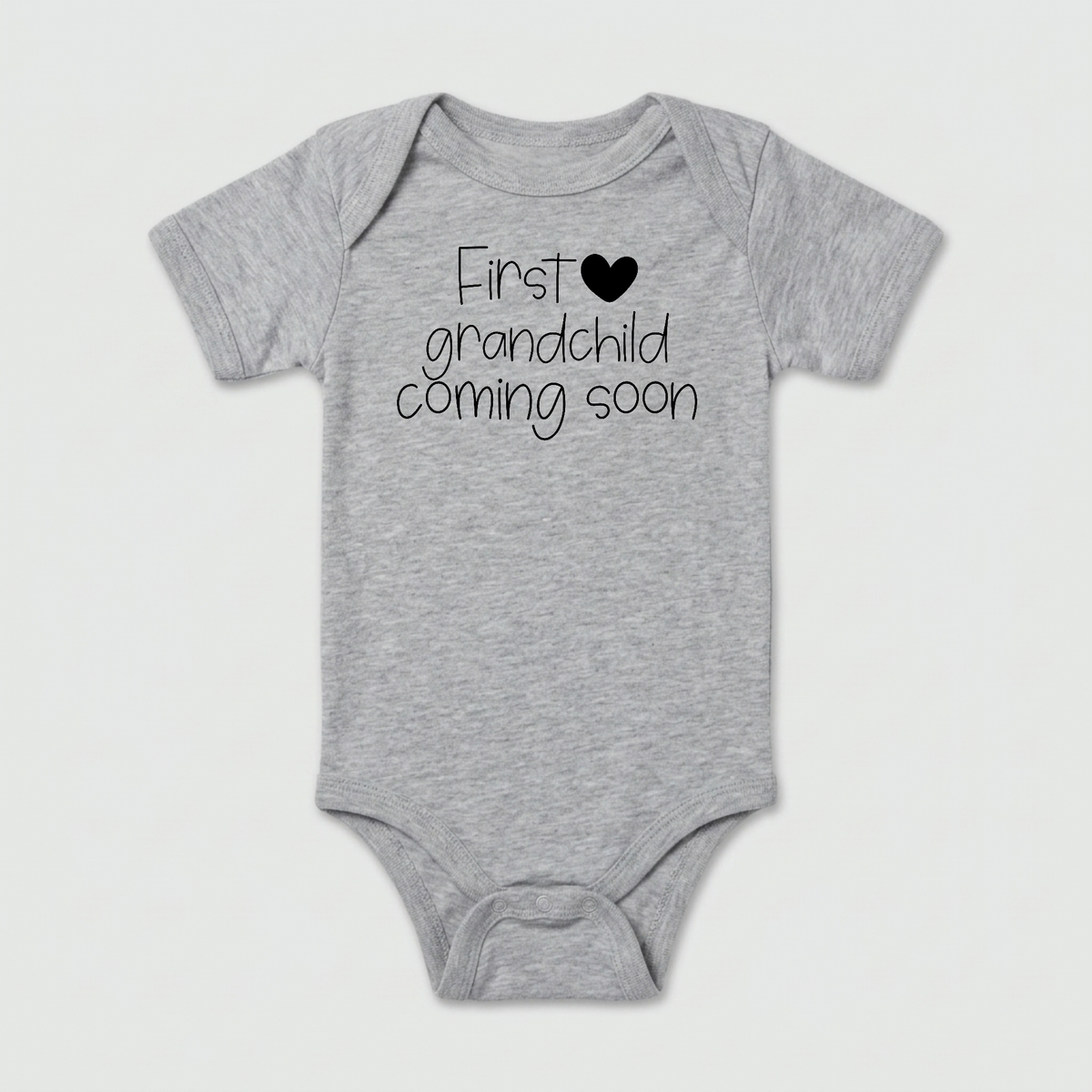 Introducing the First Grandchild Coming Soon Baby Onesie – Grandparent Announcement: a gray baby bodysuit featuring “First grandchild coming soon” in black lettering with a black heart above “grandchild.” Perfect for pregnancy announcements or celebrating your special first grandchild.