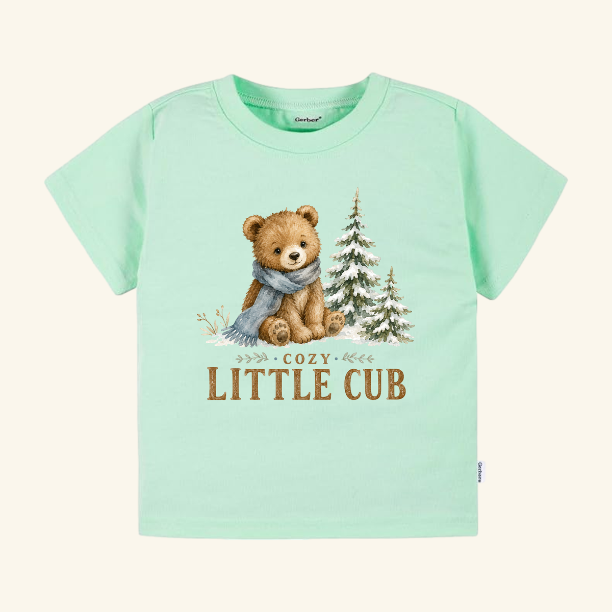 Light green Cozy Little Cub Toddler Shirt – Cute Winter Bear Tee featuring a bear wearing a blue scarf next to two snow-dusted pine trees.