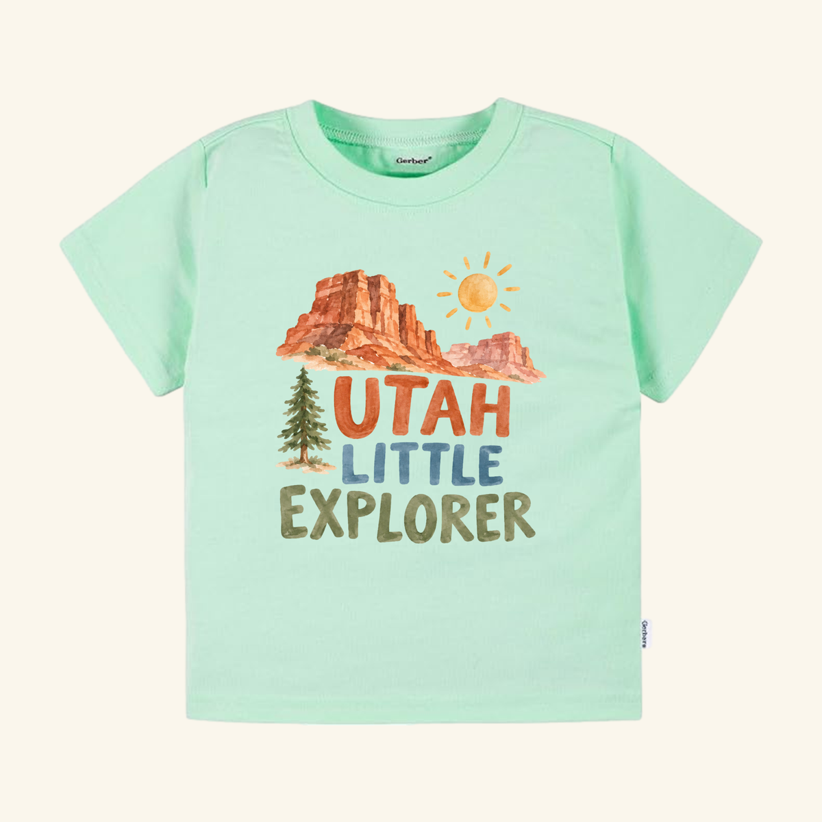 Light green Utah Toddler Shirt – Little Explorer Red Rock Desert Tee featuring a watercolor graphic of red rock desert landscapes, a pine tree, a sun, and the text UTAH LITTLE EXPLORER.