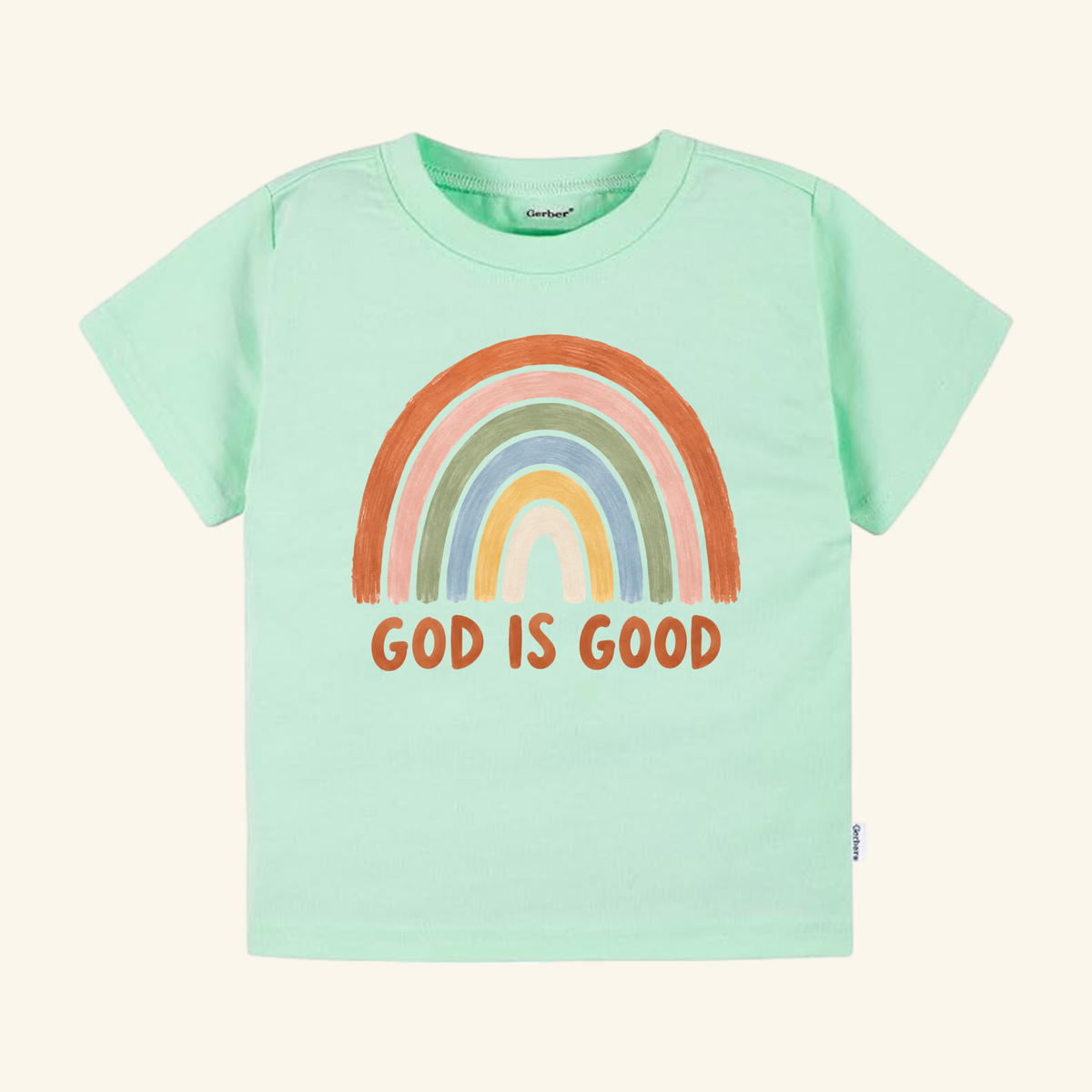 Toodleroos Christian Toddler Shirt – God Is Good Rainbow Tee: light green shirt with a hand-painted orange, gray, yellow, and beige rainbow and bold GOD IS GOOD text.