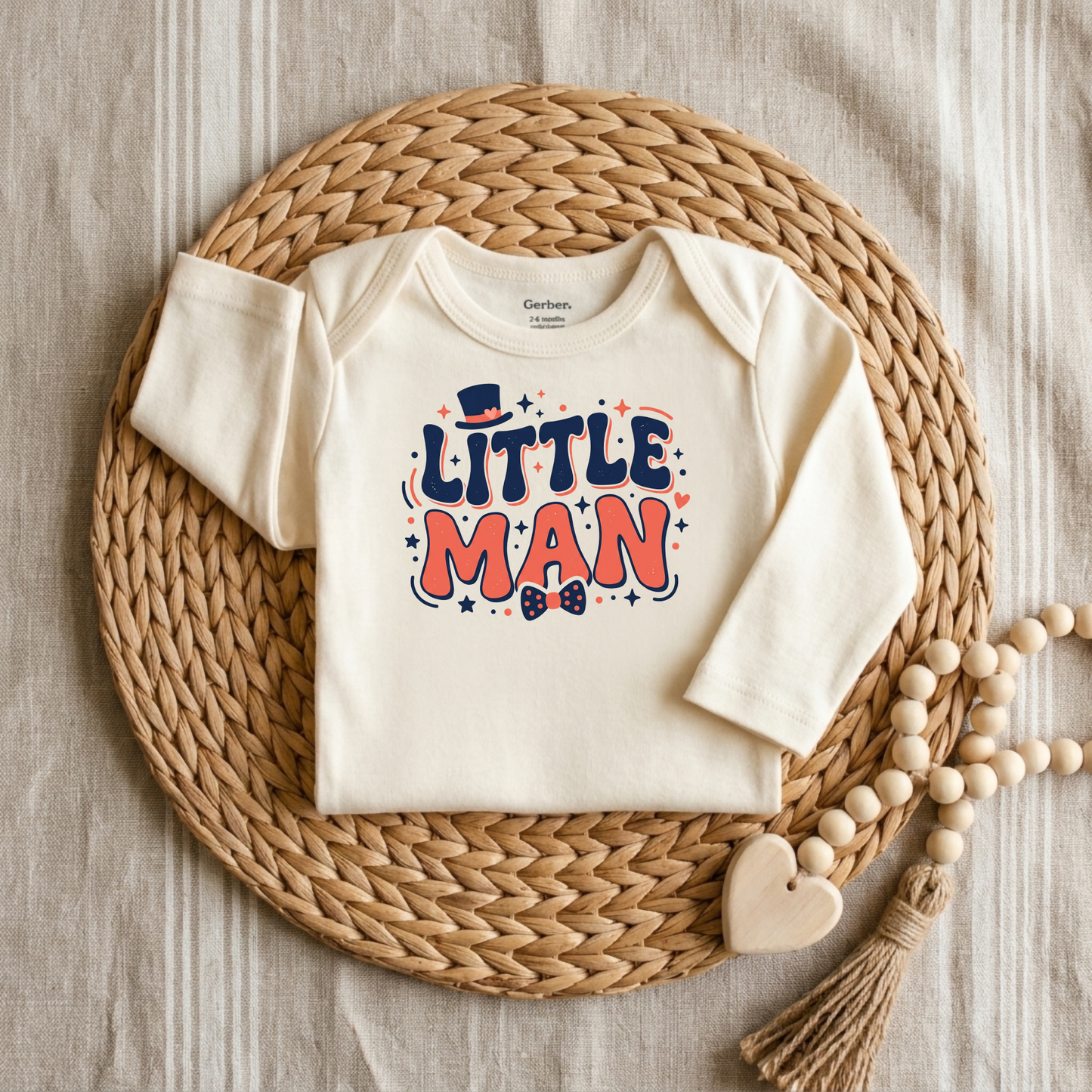 An off-white long-sleeve baby bodysuit with a colorful 'LITTLE MAN' graphic, styled flat-lay on a round woven straw mat with wooden beads on a neutral fabric background.