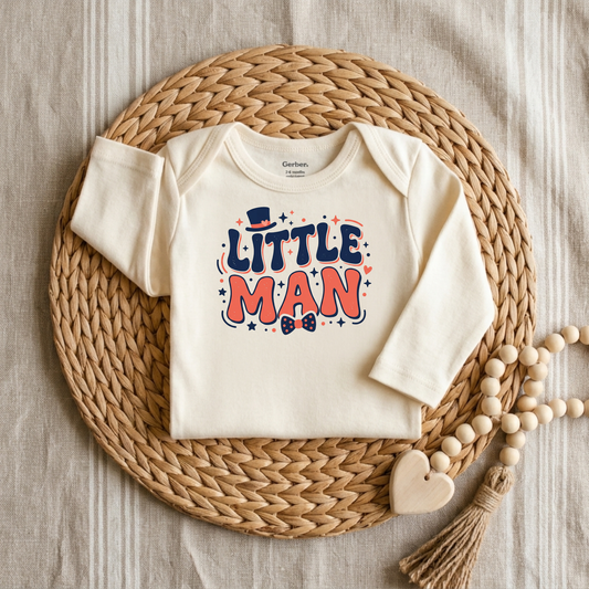 An off-white long-sleeve baby bodysuit with a colorful 'LITTLE MAN' graphic, styled flat-lay on a round woven straw mat with wooden beads on a neutral fabric background.
