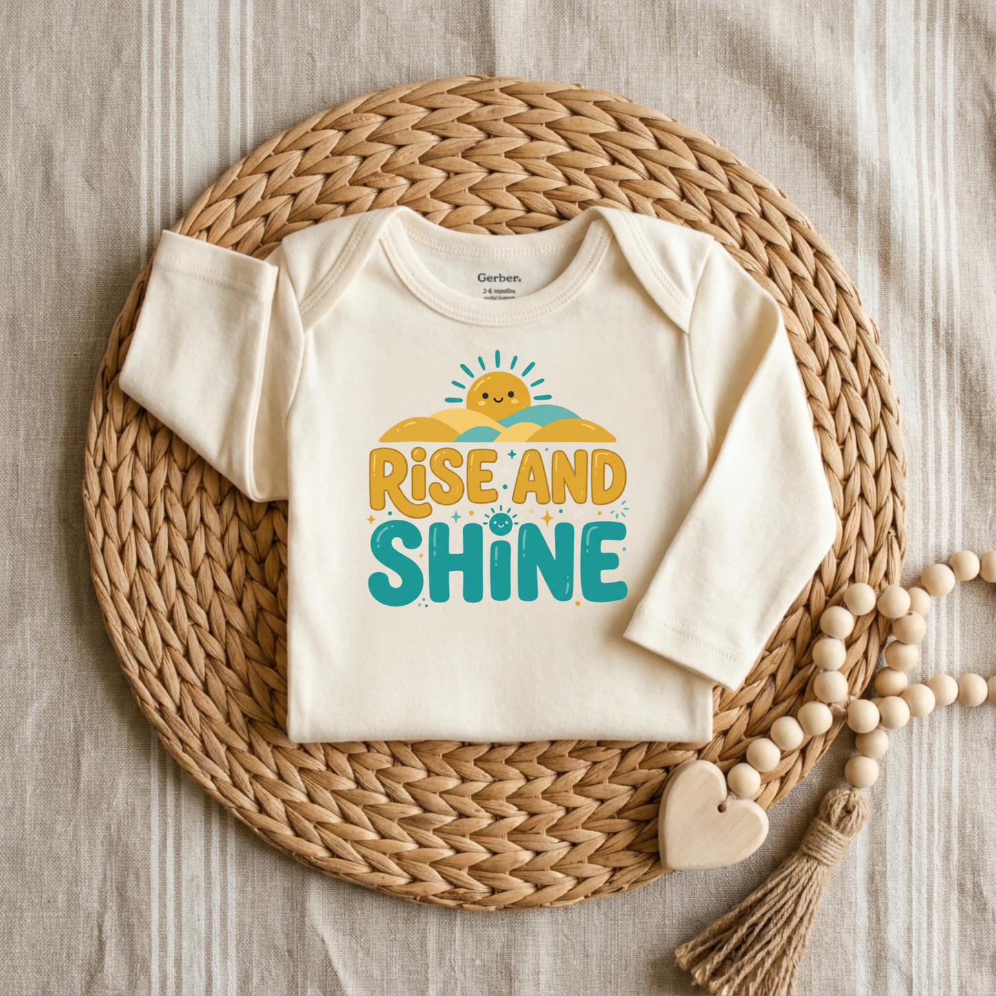 An off-white long-sleeve baby bodysuit with a colorful 'RISE AND SHINE' graphic, styled flat-lay on a round woven straw mat with wooden beads on a neutral fabric background.