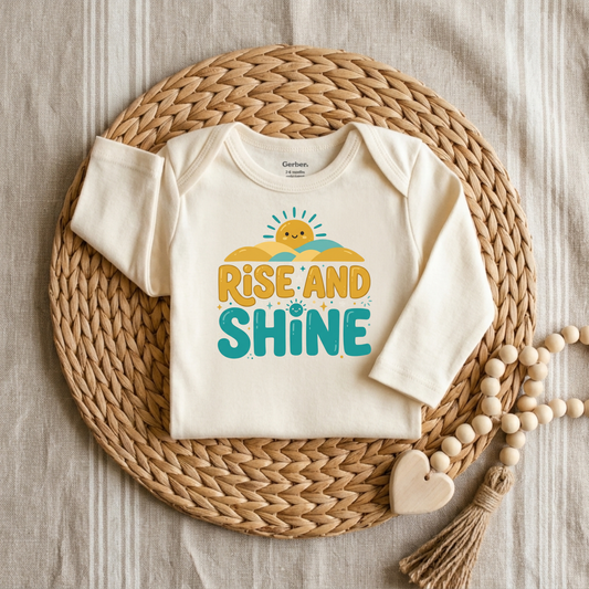 An off-white long-sleeve baby bodysuit with a colorful 'RISE AND SHINE' graphic, styled flat-lay on a round woven straw mat with wooden beads on a neutral fabric background.