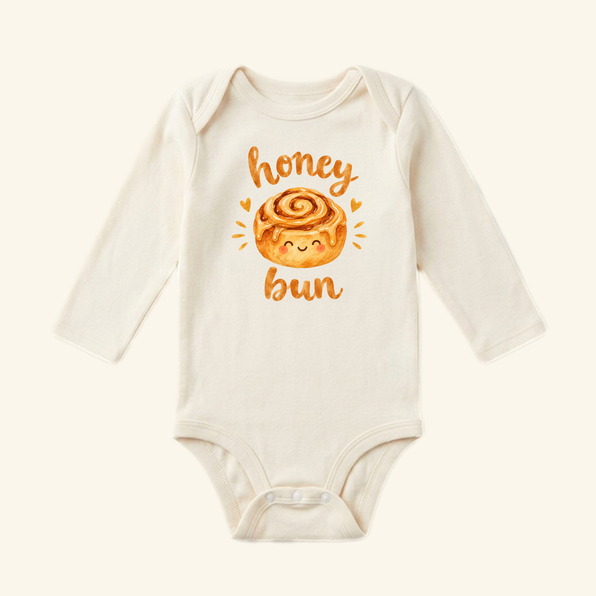 The Toodleroos Cute Baby Onesie – Honey Bun Cinnamon Roll Bodysuit features a smiling cinnamon roll with honey bun in golden letters—a sweet, cream-colored baby outfit.