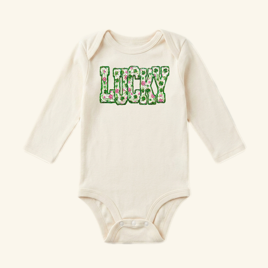 Toodleroos St. Patrick’s Day Baby Onesie – Lucky Floral Clover Bodysuit in cream with long sleeves and the word LUCKY printed in a green clover and pink floral pattern across the chest.