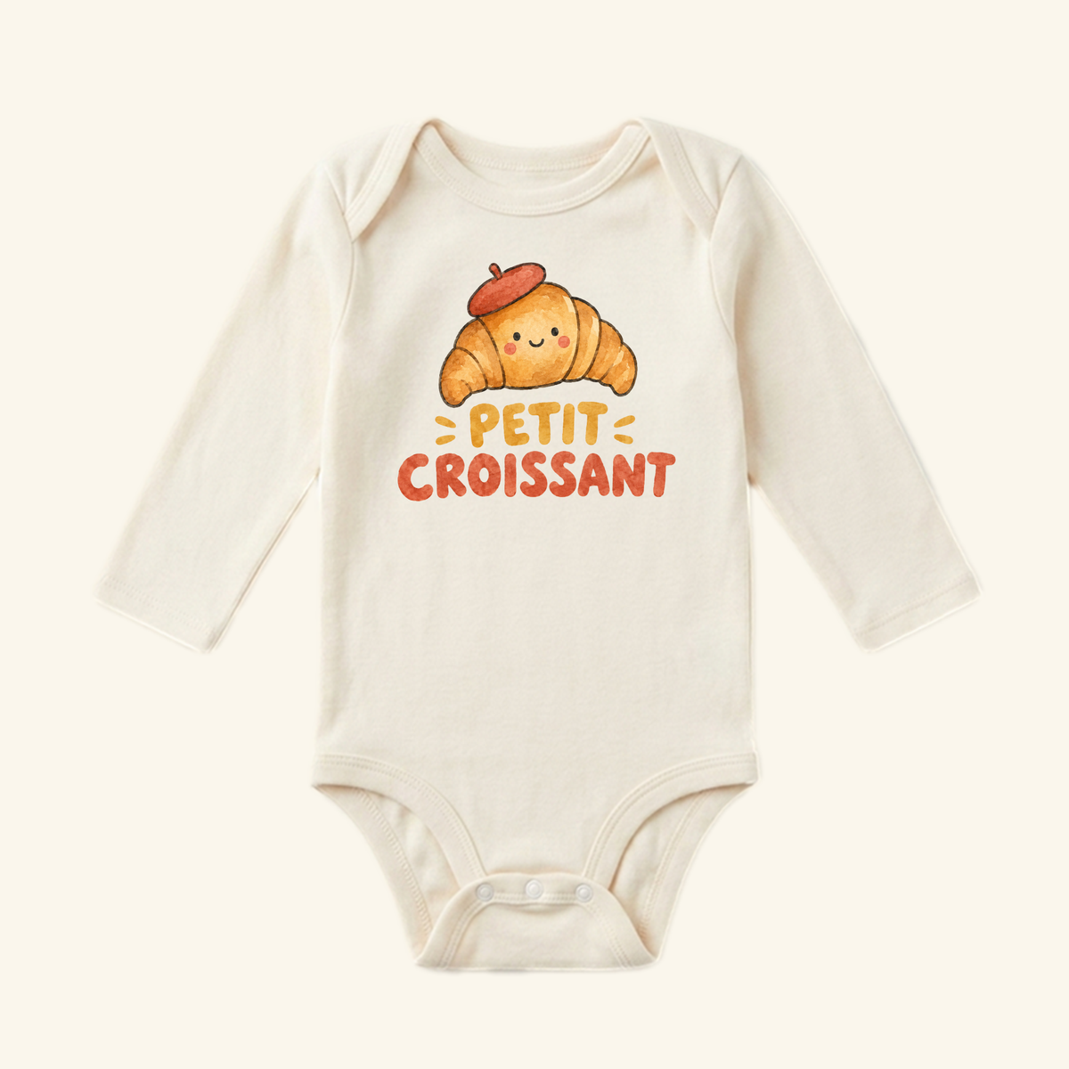 Cream long-sleeved Toodleroos Petit Croissant Baby Onesie featuring a graphic of a smiling croissant wearing a red beret with the text PETIT CROISSANT underneath.