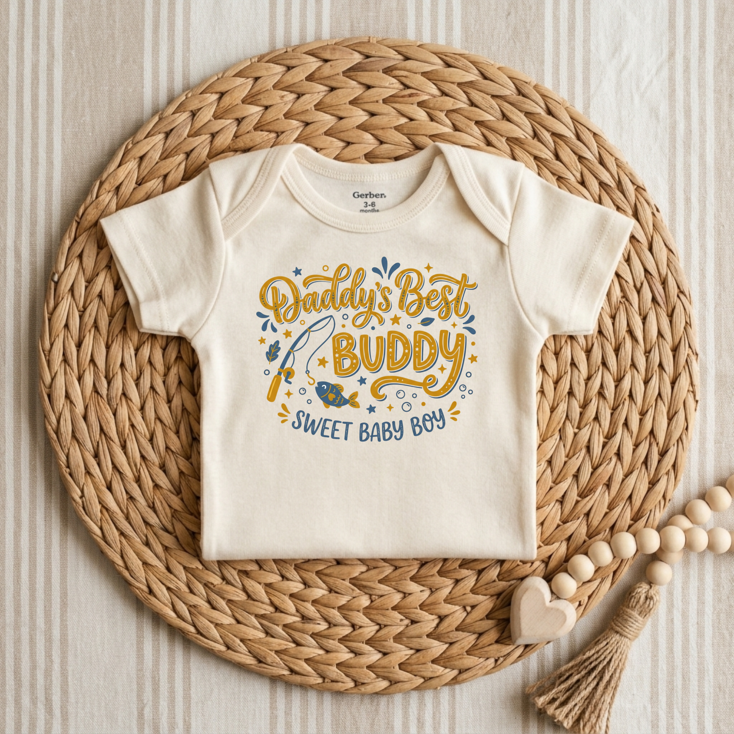 An off-white short-sleeve baby bodysuit with a colorful graphic reading 'Daddy's Best Buddy Sweet Baby Boy' in yellow and blue, styled flat on a round woven straw mat with wooden beads and a tassel.