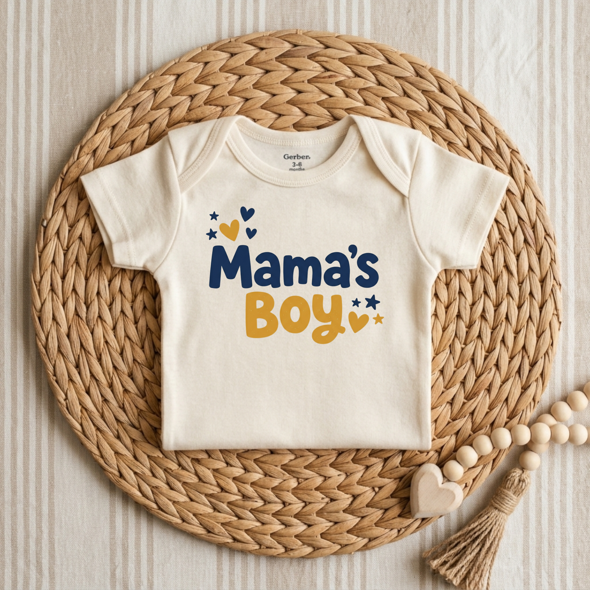 An off-white short-sleeve baby bodysuit with 'Mama's Boy' printed in navy and mustard yellow, styled on a round woven straw mat with wooden beads.