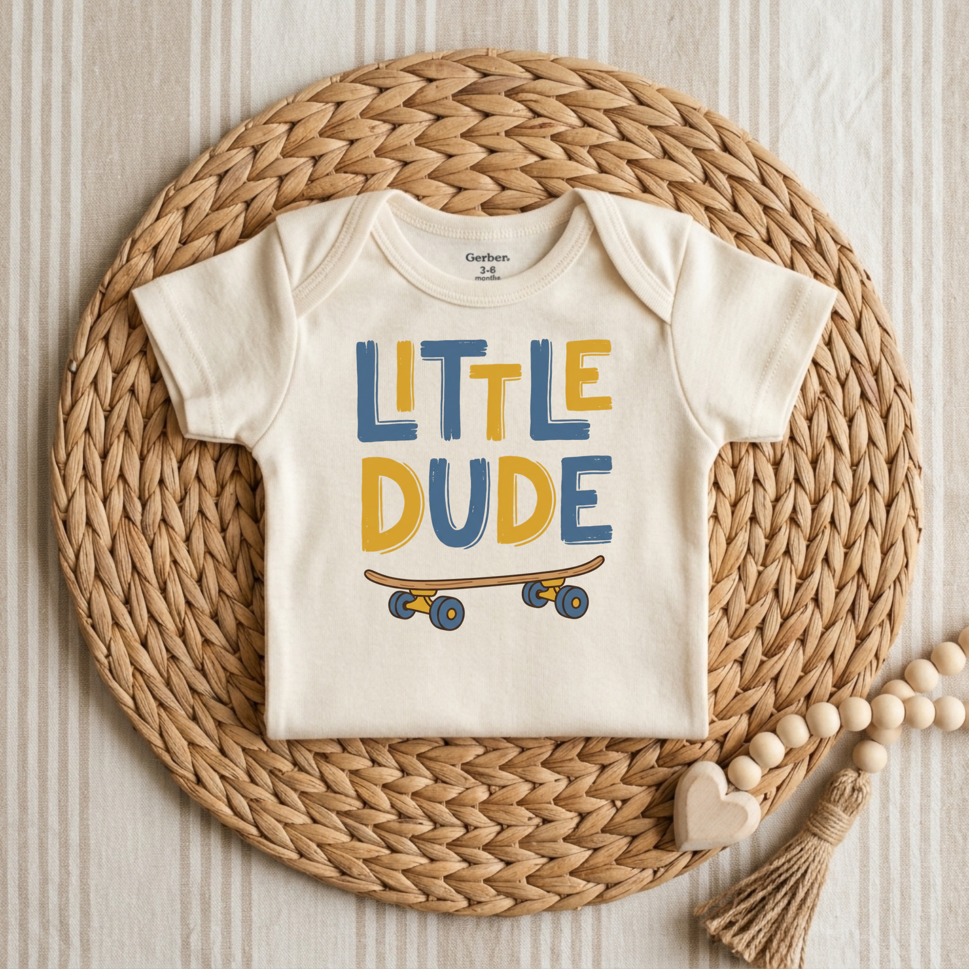 An off-white short-sleeve baby bodysuit with 'LITTLE DUDE' and a skateboard graphic in blue and mustard yellow, styled flat on a round woven straw mat.