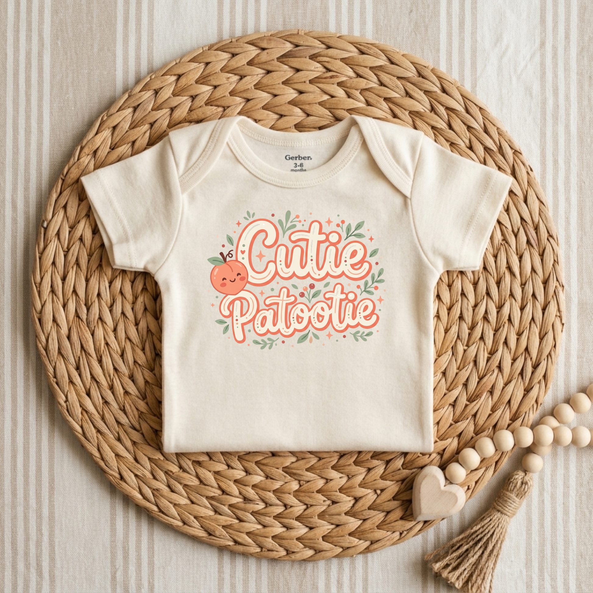 An off-white short-sleeve baby bodysuit with a 'Cutie Patochie' graphic, styled flat-lay on a round woven straw mat with wooden beads and a heart tassel.