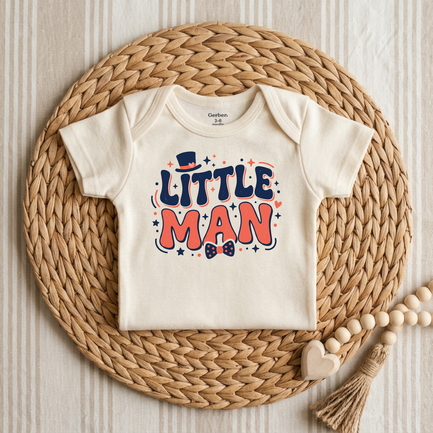 An off-white short-sleeve baby bodysuit with a colorful 'LITTLE MAN' graphic print, styled flat on a round woven straw mat with wooden beads and a heart tassel.