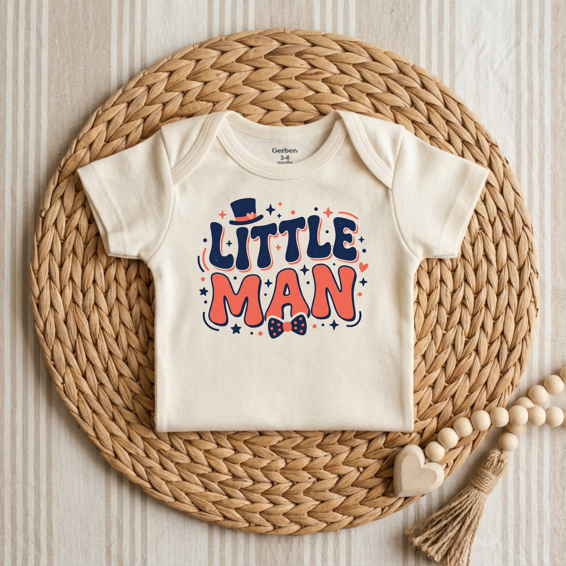 An off-white short-sleeve baby bodysuit with a colorful 'LITTLE MAN' graphic print, styled flat on a round woven straw mat with wooden beads and a heart tassel.