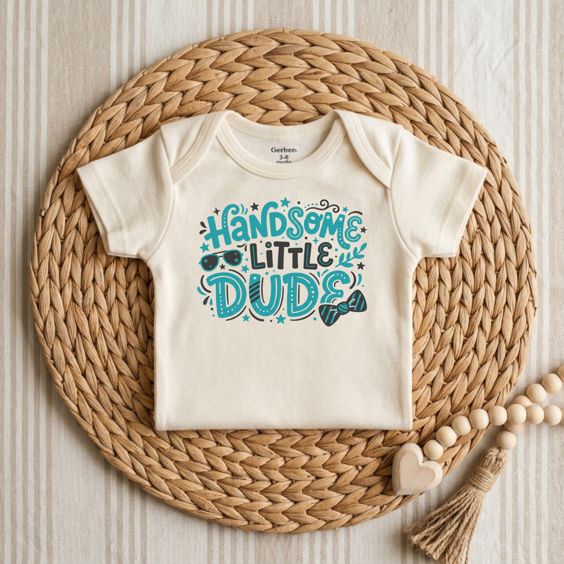 An off-white short-sleeve baby bodysuit with a colorful graphic reading 'HANDSOME LITTLE DUDE' in teal and black, styled flat on a round woven straw mat with wooden beads and a tassel.