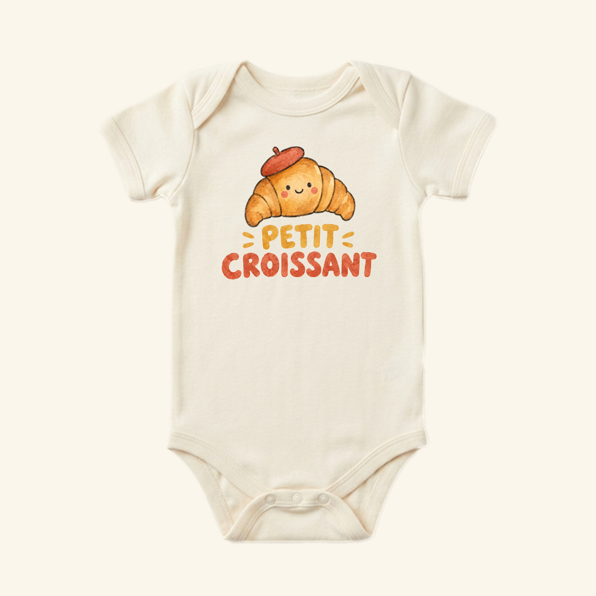 Cream-colored Petit Croissant Baby Onesie featuring an illustration of a smiling croissant wearing a beret, with the text Petit Croissant printed below.