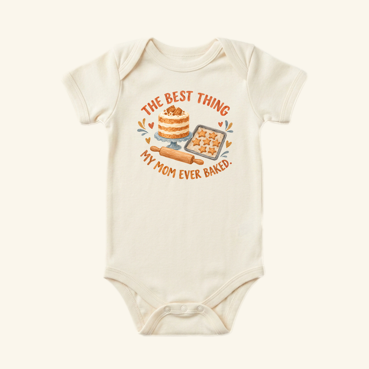 Off-white Best Thing My Mom Baked Baby Onesie – Cute Mom Baking Bodysuit featuring a graphic of a layer cake, star-shaped cookies, and a rolling pin with the caption THE BEST THING MY MOM EVER BAKED.