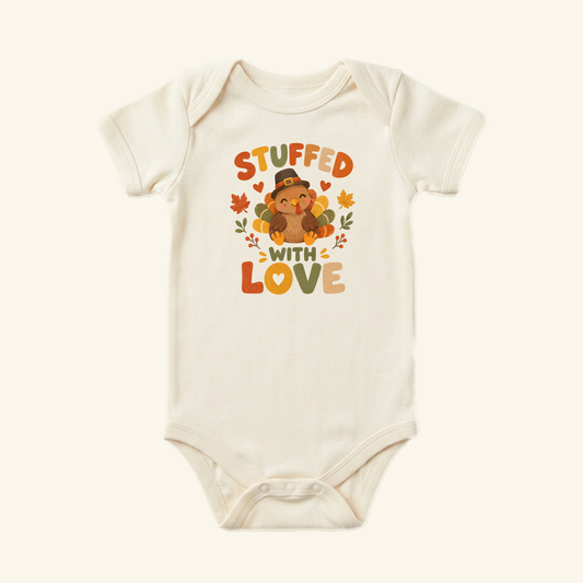 Cream-colored Stuffed With Love Baby Onesie – Thanksgiving Turkey Bodysuit featuring a cartoon turkey in a pilgrim hat, autumn leaves, and the text Stuffed With Love.
