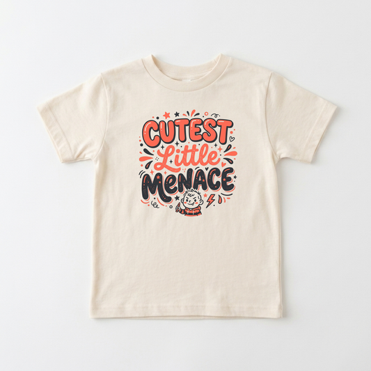 A cream-colored short-sleeve t-shirt with a colorful graphic on the chest that reads 'CUTEST Little MENACE' in orange, black, and pink lettering, accompanied by a small cartoon character.