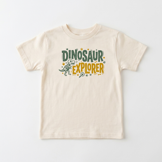 A light beige short-sleeve t-shirt with a 'Dinosaur Explorer' graphic on the chest, featuring a small dinosaur illustration and colorful text, displayed on a clean white background.