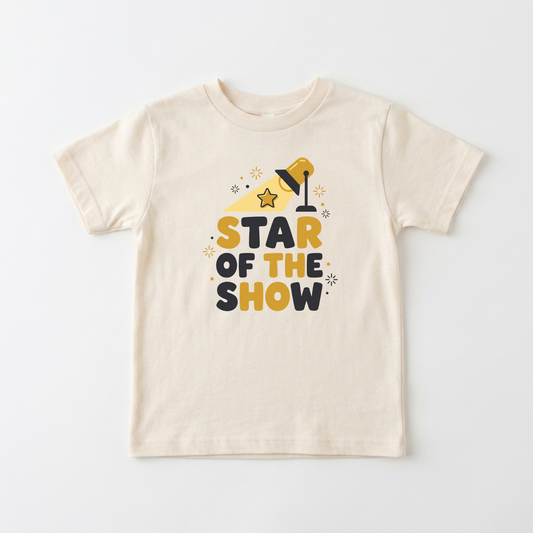A cream-colored short-sleeve kids' t-shirt with a graphic print that reads 'STAR OF THE SHOW' in yellow and dark navy blue letters, accompanied by a yellow spotlight and star illustrations, displayed on a clean white background.