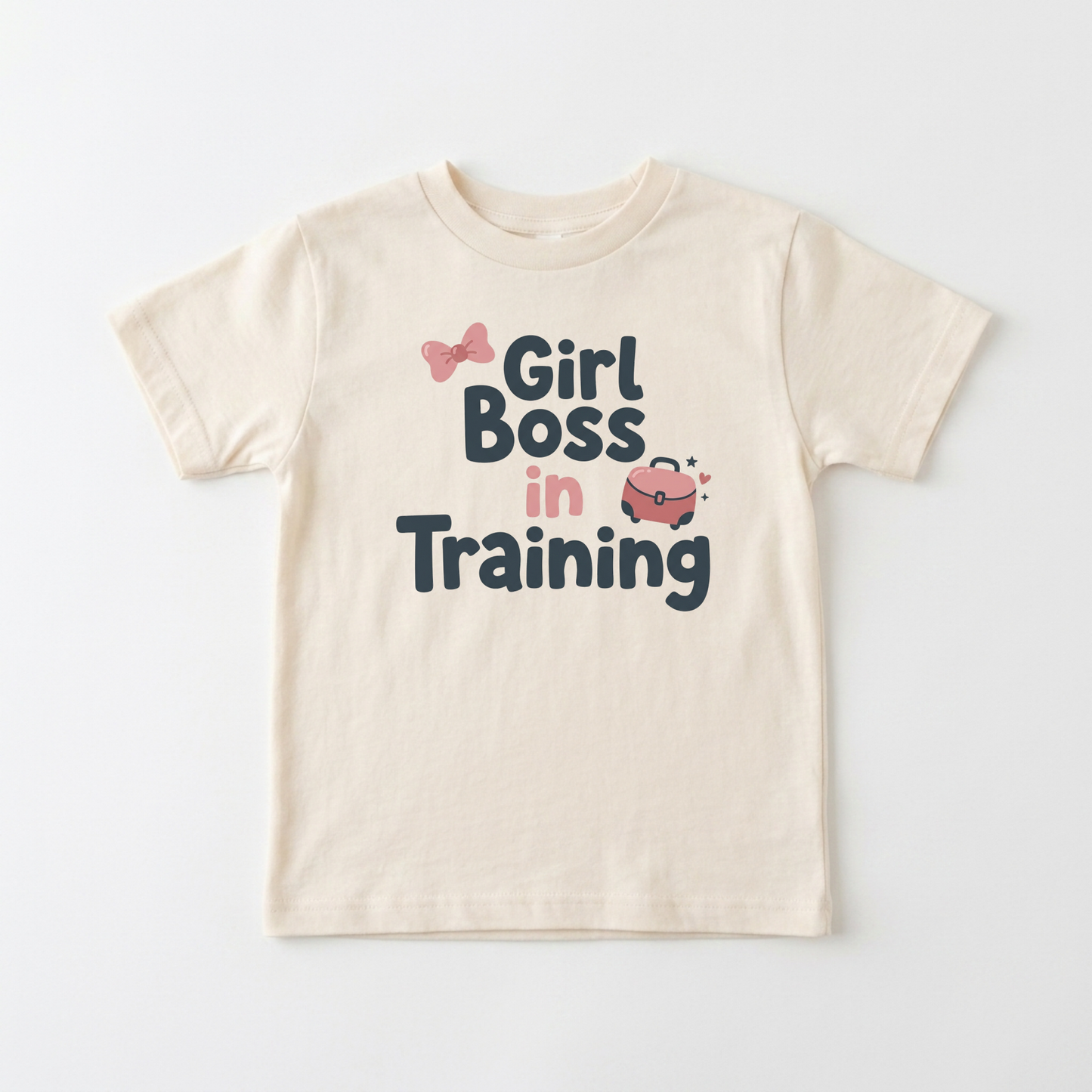 A cream-colored short-sleeve t-shirt with a graphic print that reads 'Girl Boss in Training' in dark blue and pink lettering, accompanied by small pink bow and purse illustrations, shown on a light gray background.