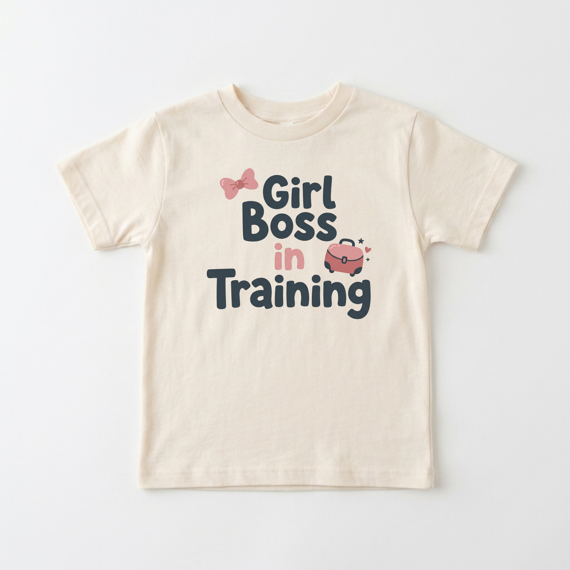 A cream-colored short-sleeve t-shirt with a graphic print that reads 'Girl Boss in Training' in dark blue and pink lettering, accompanied by small pink bow and purse illustrations, shown on a light gray background.