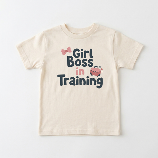 A cream-colored short-sleeve t-shirt with a graphic print that reads 'Girl Boss in Training' in dark blue and pink lettering, accompanied by small pink bow and purse illustrations, shown on a light gray background.