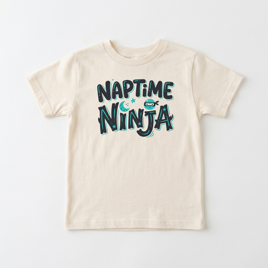 A cream-colored short-sleeve kids' t-shirt with a 'NAPTIME NINJA' graphic printed on the chest in dark teal and light blue, laid flat against a light gray background.
