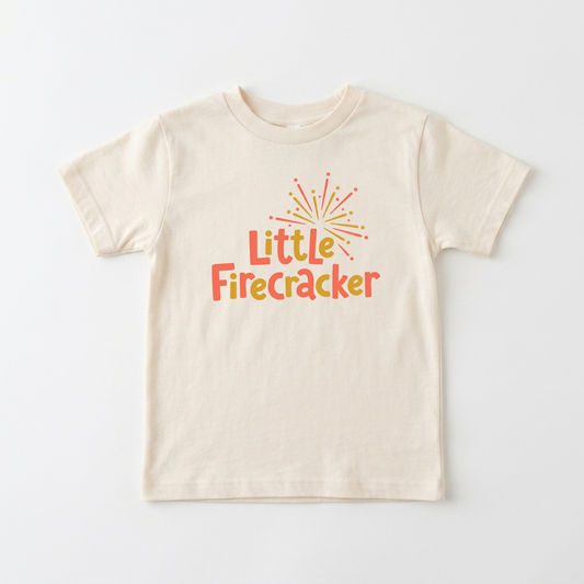 A cream-colored short-sleeve t-shirt with a colorful graphic on the chest that reads 'Little Firecracker' in orange and yellow text under a small firework illustration, displayed flat against a light gray background.