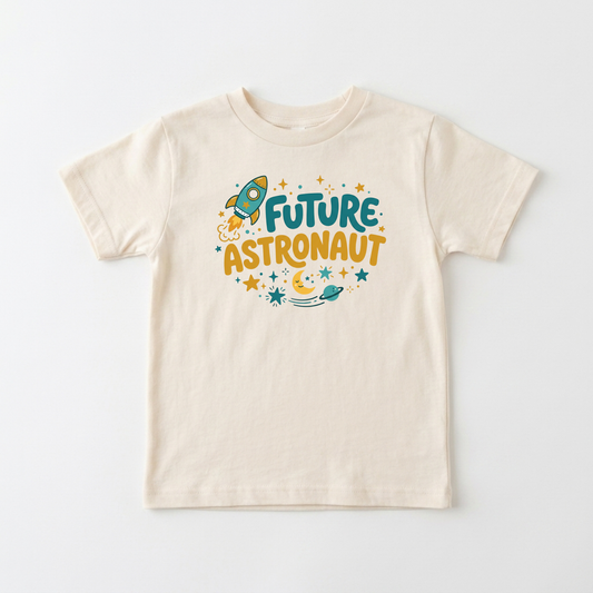 A cream-colored short-sleeve t-shirt with a colorful graphic on the chest that reads 'FUTURE ASTRONAUT' in teal and mustard yellow, surrounded by a rocket ship, stars, and a crescent moon, displayed on a clean white background.