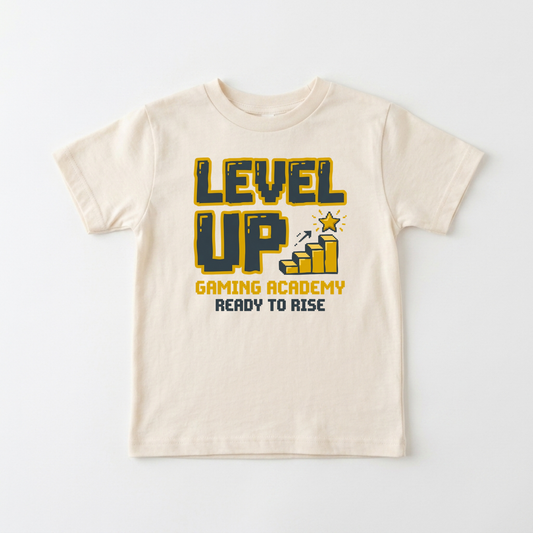 A cream-colored short-sleeve t-shirt with a graphic on the front that reads 'LEVEL UP GAMING ACADEMY READY TO RISE' in yellow and dark blue, accompanied by a small bar graph and star icon.