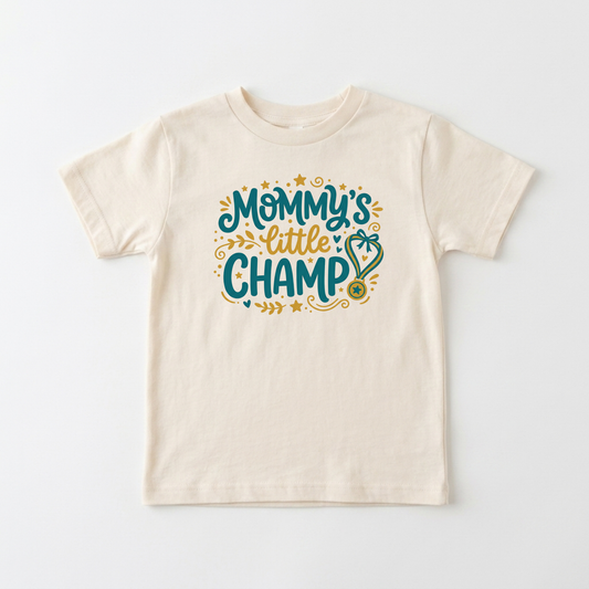An off-white short-sleeve kids' t-shirt with a colorful graphic on the chest that reads 'Mommy's little CHAMP' in teal and gold lettering with decorative stars and a stethoscope.