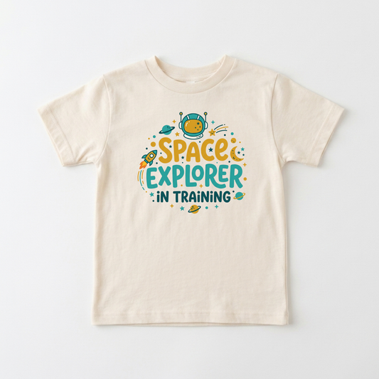 A cream-colored short-sleeve t-shirt with a colorful graphic on the front that reads 'SPACE EXPLORER IN TRAINING' in yellow and teal lettering with cartoon space elements, shown on a light gray background.