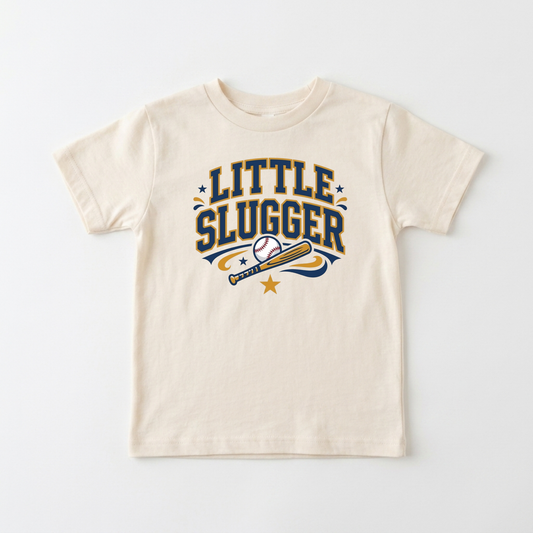 A cream-colored short-sleeve t-shirt with a 'LITTLE SLUGGER' baseball graphic in navy blue and gold, displayed flat against a light gray background.