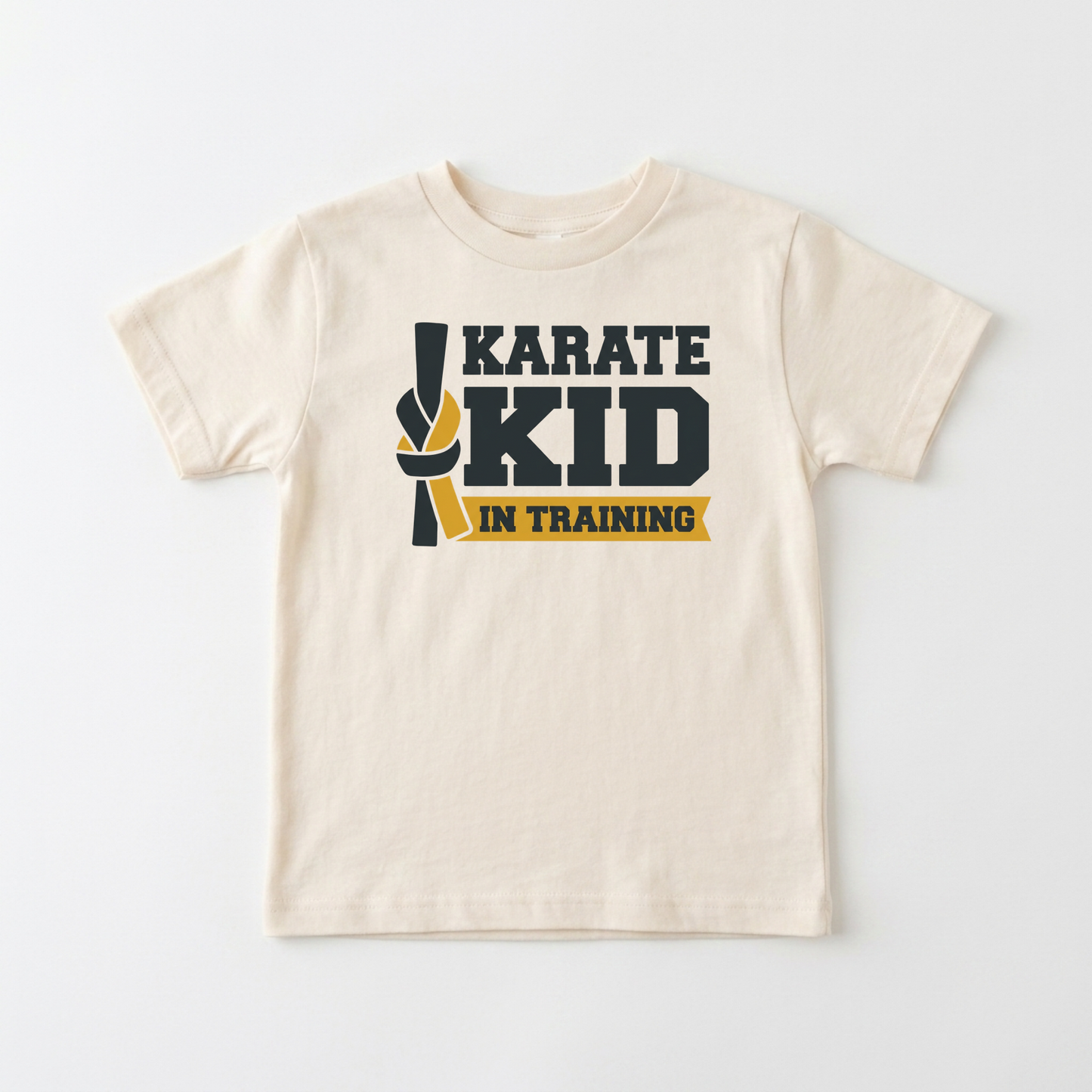 A cream-colored short-sleeve kids' t-shirt with a 'KARATE KID IN TRAINING' graphic on the chest, featuring a black and yellow martial arts ribbon design, laid flat against a light gray background.